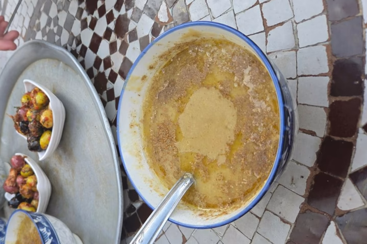 A bowl of bissara with cumin and olive oil beside olives on a zellige table in Tangier